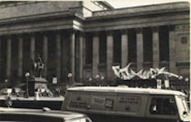 Large inflatable sculpture in front of civic building with a crowd of people and street traffic