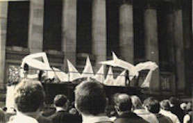 Large inflatable sculpture in front of civic building with a crowd of people and street traffic