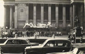 Large inflatable sculpture in front of civic building with a crowd of people and street traffic